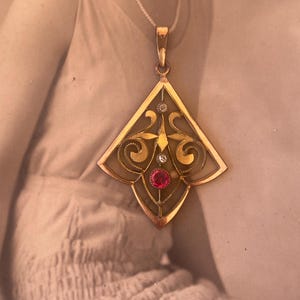 May include: A gold pendant with a geometric design featuring a red ruby and two smaller diamonds. The pendant is in the shape of a diamond and has intricate filigree details.