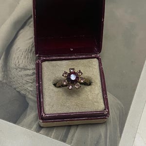 Puede incluir: Un anillo de tono dorado con una piedra central de color rojo oscuro rodeada por seis piedras más pequeñas y claras en forma de flor, presentado dentro de una pequeña caja cuadrada de color granate con forro interior de terciopelo beige.