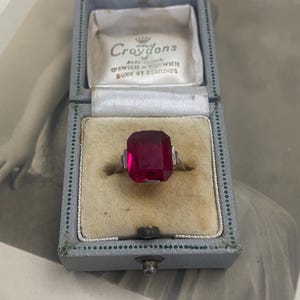 May include: A vintage ring with a large, rectangular, ruby-red gemstone set in a gold band. The ring is displayed in a light blue box with the name "Croydons" printed on the inside lid.