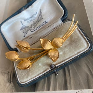 May include: A gold-toned metal brooch shaped like a floral spray with three closed tulip-like buds and one open three-petaled flower, resting inside a dark blue velvet-lined jewellery box with the text "Watchmaker & Jeweller Robert Crawford Cumnock".