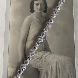 Può includere: Un braccialetto vintage in argento con pietre chiare e scintillanti. Il braccialetto presenta un design intricato di forme alternate ed è esposto su una fotografia color seppia di una donna.