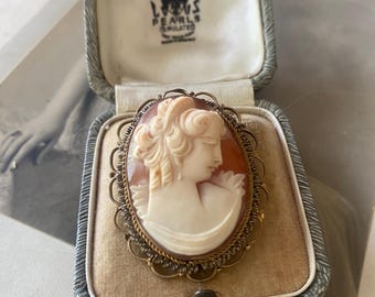 May include: A cameo brooch with a gold-tone filigree setting. The cameo features a carved portrait of a woman with a delicate floral design. The brooch is in a small, gray velvet box with the inscription "Lewis Pearls Simulated" on the inside of the lid.