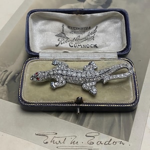 May include: A silver lizard brooch with red eyes, set with rhinestones, in a velvet-lined box. The box is labelled "Watchmakers & Jeweller Robert Crawford Cumnock."