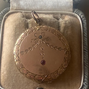 Könnte beinhalten: Ein goldenes Medaillon mit einem zarten floralen Design und einem roten Edelstein-Akzent. Das Medaillon befindet sich in einer samtenen Box.