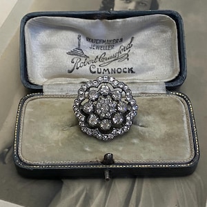 Puede incluir: Un anillo tipo cluster de tono plateado con una disposición central de piedras redondas transparentes rodeadas por un borde festoneado de piedras más pequeñas, presentado en un joyero rectangular azul oscuro con el texto Robert Crawford Watchmaker & Jeweller Cumnock.