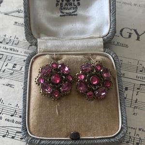 Könnte beinhalten: Ein Paar Vintage-Ohrringe mit rosa Strasssteinen in einem silberfarbenen Etui. Die Ohrringe, gefasst in goldfarbenem Metall, weisen mehrere rosa Steine auf. Das offene Etui zeigt die Ohrringe auf einem beigefarbenen Samtfutter.