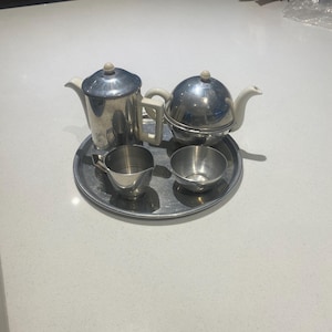 Puede incluir: Un juego de té de plata con una tetera, una jarra de crema y dos azucareros. La tetera tiene un cuerpo redondo y una tapa blanca. La jarra de crema y los azucareros son redondos y tienen un acabado plateado.