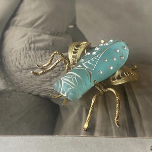 May include: A detailed, vintage-style insect brooch. The body is a light blue, possibly glass, with etched details and small clear stones. The wings and legs are gold-toned metal, creating a striking contrast.