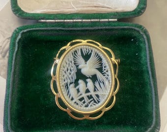 May include: A gold-coloured brooch with a white cameo depicting birds in a tree, set against a black background. The brooch is presented in a green velvet jewellery box with a gold clasp. The cameo features a detailed relief carving.