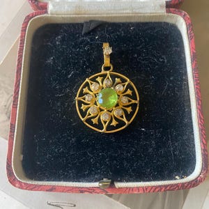 Puede incluir: Colgante de oro con una piedra preciosa verde central, rodeada de piedras claras más pequeñas y un diseño de filigrana dorada decorativa. El colgante se muestra en un joyero rojo y negro, lo que sugiere una pieza vintage o antigua.