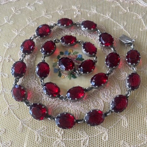 Pode incluir: Um conjunto de joias vintage com colar e pulseira. Ambas as peças apresentam pedras preciosas ovais, vermelho-escuras, em metal prateado. O colar e a pulseira são exibidos em uma superfície creme, semelhante a renda.