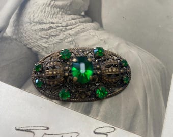 May include: A vintage brooch with a filigree design and green glass stones. The brooch is oval shaped and has a central green stone surrounded by smaller green stones.