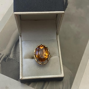 May include: A vintage gold ring featuring a large, oval amber gemstone, set in an ornate filigree design. The ring is presented in a grey and white ring box, on a vintage photograph.