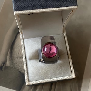 Könnte beinhalten: Ein silberfarbener Ring mit einem großen, runden, rosa Edelstein in einem rechteckigen Rahmen. Der Ring wird in einer Vintage-Schmuckschatulle mit dunkelblauem, strukturiertem Äußeren und cremefarbenem Innenraum präsentiert.