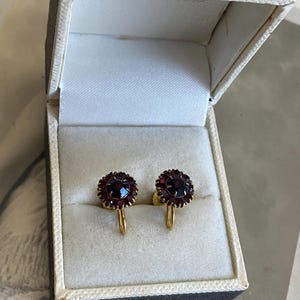 Puede incluir: Un par de pendientes redondos de tornillo con una piedra preciosa roja oscura central rodeada por un halo de piedras rojas más pequeñas, engastadas en metal dorado. Los pendientes se presentan dentro de un joyero rectangular con forro blanco.