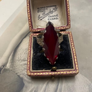 May include: Antique ring featuring a large, faceted, deep red gemstone in an ornate setting. The ring is presented in a vintage jewelry box. The box has the text "Geo Beasley & Co, Jewellers & Opticians, Cornhill, Chard."