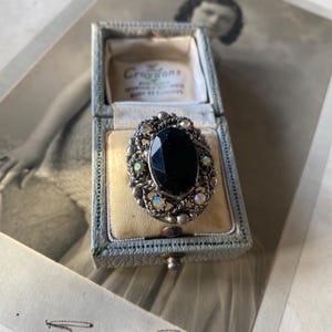 Peut inclure: Bague ancienne ornée d'une grande pierre noire ovale sertie dans un cadre en métal argenté. Le cadre est orné de petites pierres iridescentes. La bague est présentée dans un écrin bleu vintage, sur une photographie ancienne.