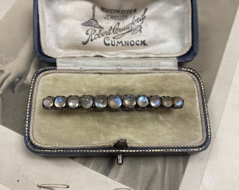 Könnte beinhalten: Eine antike Spangenbrosche mit einer Reihe schillernder Mondstein-Cabochons, gefasst in silberfarbenem Metall. Die Brosche wird in einer blau samtgefütterten Schatulle mit dem Text "Robert Crawford, Watchmaker & Jeweller, Cumnock" präsentiert.