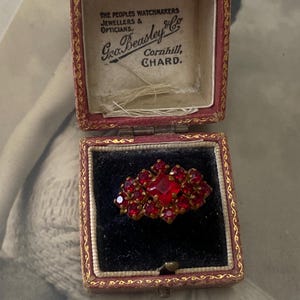 Op de afbeelding: Vintage ring in een rood en goudkleurig doosje. De ring heeft een centrale vierkante rode steen, omringd door kleinere rode stenen. Het doosje is rood met gouden randen en de tekst "Gro. Beasley & Co" is zichtbaar.