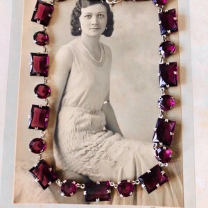 May include: A vintage necklace with a silver chain and large, faceted, purple glass stones. The necklace is displayed on a black and white photograph of a woman.