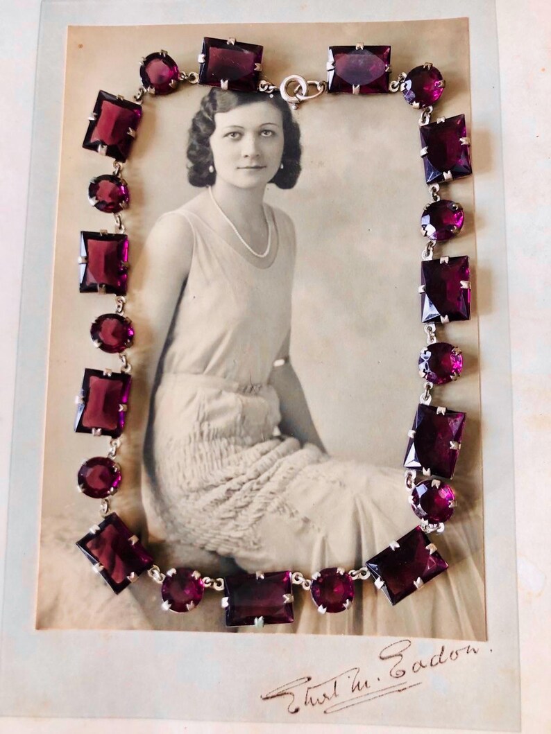 May include: A vintage necklace with a silver chain and large, faceted, purple glass stones. The necklace is displayed on a black and white photograph of a woman.