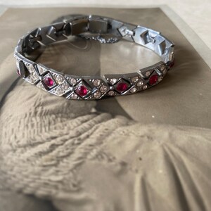 Pode incluir: Uma pulseira prateada com pedras preciosas vermelhas e transparentes alternadas. A pulseira está sobre uma fotografia vintage. As pedras preciosas estão dispostas em um padrão geométrico, criando um acessório brilhante e elegante.