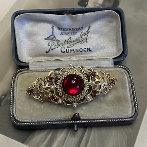 May include: A gold-tone filigree brooch featuring a large central red cabochon, smaller red stones, and tiny white pearls. The brooch is displayed inside a dark blue hinged jewellery box with the text "WATCHMAKERS JEWELLER Robert Crawford CUMNOCK".