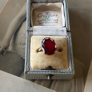 Puede incluir: Un anillo vintage con una gran gema roja oscura ovalada, engastada en una banda plateada. Se muestra en un joyero azul claro abierto, con el nombre "Croydons" impreso en el interior. El joyero está forrado con terciopelo crema.