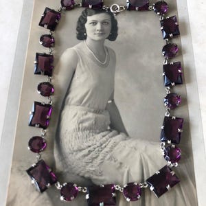 May include: A vintage silver necklace with a chain and purple square and round glass stones. The necklace is displayed on a black and white photograph of a woman.