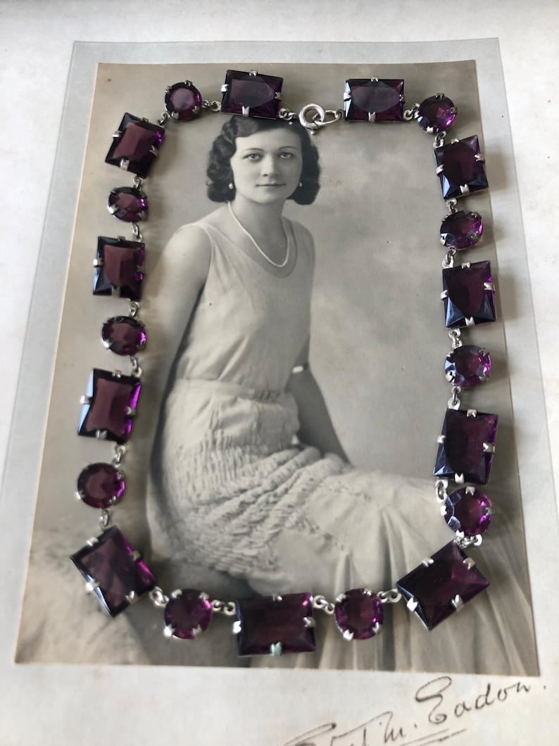 May include: A vintage silver necklace with a chain and purple square and round glass stones. The necklace is displayed on a black and white photograph of a woman.