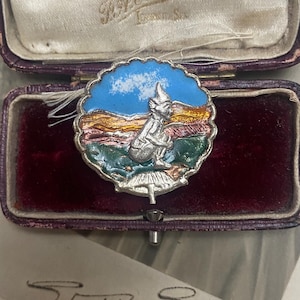 Könnte beinhalten: Eine silberne und emaillierte Anstecknadel mit einem fantasievollen Kobold, der auf einem Pilz sitzt. Der Kobold trägt einen spitzen Hut und hat einen blauen und weißen Himmel hinter sich. Die Anstecknadel befindet sich in einer roten Samtbox mit einem weißen Etikett, auf dem "T.H. Eaton, London." steht.