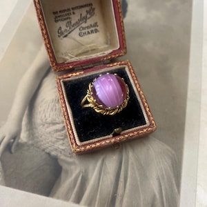 May include: A vintage ring with a large, oval, striped, pink gemstone set in a gold-coloured metal band. The ring is displayed in a red and gold jewellery box with the text "Geo. Beasley & Co." The box is on top of a vintage photograph.