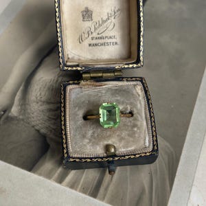 May include: A gold ring featuring a rectangular, light green faceted gemstone, presented in a dark blue velvet-lined jewellery box. The interior lid of the box displays the text W.B. Riddiough & Son, St Ann's Place, Manchester, with a crown emblem.