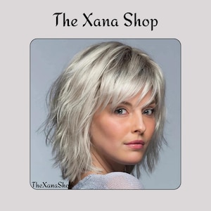 Puede incluir: Una mujer con cabello corto, plateado y en capas, con flequillo. La imagen está enmarcada con un borde gris claro y presenta el texto "The Xana Shop" en la parte superior y "TheXanaShop" en la parte inferior.