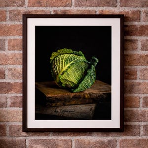 Könnte beinhalten: Gerahmtes Foto eines grünen Kohls mit strukturierten Blättern. Der Kohl liegt auf einer verwitterten Holzoberfläche vor einem schwarzen Hintergrund. Der Rahmen ist dunkelbraun, der Hintergrund eine Backsteinmauer.