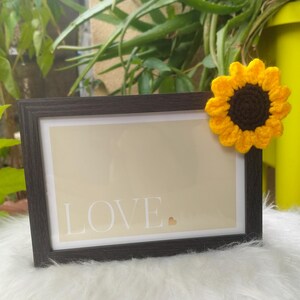 Peut inclure: Un cadre photo rectangulaire marron foncé contenant un insert beige avec le texte blanc LOVE et un petit symbole de cœur. Un tournesol au crochet jaune vif et marron foncé est fixé sur le coin supérieur droit du cadre.