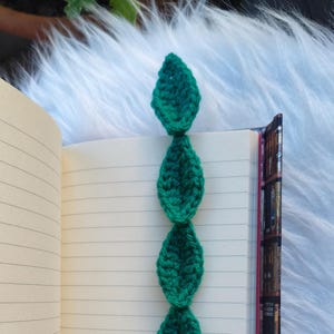Peut inclure: Marque-page fait à la main en crochet vert en forme de feuilles. Le marque-page est placé dans un cahier ouvert avec des pages lignées. Le fond est un matériau doux et blanc.