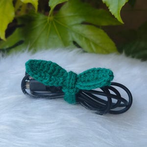 May include: A handmade, green crocheted cable tie, shaped like a bow, neatly secures a black charging cable. The tie is made of a textured yarn and rests on a white, fluffy surface, with green leaves in the background.
