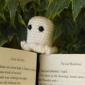 Peut inclure: Marque-page fantôme crocheté blanc avec des yeux noirs, posé sur un livre. Le titre du livre, "The Lost Bookshop", est visible. Un accessoire artisanal et original pour les lecteurs.