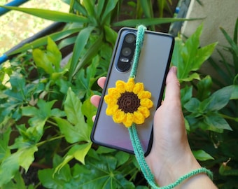 Op de afbeelding: Een grijze smartphone met een zwarte hoes en een gehaakte zonnebloem in geel en bruin, bevestigd aan een groene lanyard. De telefoon bevindt zich tegen een achtergrond van groen gebladerte en een blauwe balustrade.