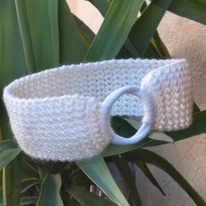 May include: White crocheted headband with a white plastic ring. The headband is lying on a green plant.