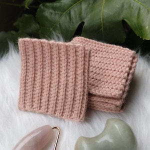 Peut inclure: Deux carrés en crochet rose, un rouleau facial en quartz rose et un outil gua sha en jade vert sur une surface blanche et moelleuse.
