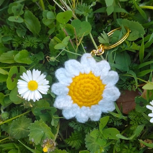 Peut inclure: Un porte-clés en forme de marguerite fait à la main, avec des pétales blancs crochetés et un centre jaune, sur fond d'herbe verte et de feuillage. Le porte-clés a un anneau doré.