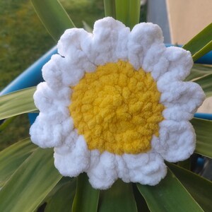 May include: A handmade crochet daisy with white petals and a yellow center. The flower is set against a backdrop of green leaves and a blue pot, creating a vibrant and cheerful display. This decorative item is perfect for home decor.