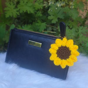 May include: Black wallet with a gold zipper and a yellow and brown crochet sunflower charm. The wallet has a gold rectangular accent. The background is a blurred green foliage and white fluffy fabric.