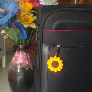 Peut inclure: Une valise noire avec un charm en forme de tournesol crocheté jaune et marron suspendu à la fermeture éclair. Un vase de fleurs artificielles colorées est à gauche de la valise.
