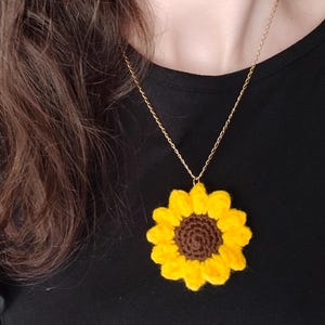 Peut inclure: Un collier pendentif en forme de tournesol crocheté, avec une chaîne dorée. Le tournesol a des pétales jaune vif et un centre marron. Le collier est porté sur un haut noir.