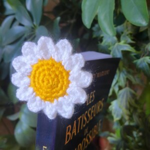 Peut inclure: Un marque-page en forme de marguerite crochetée, avec des pétales blancs et un centre jaune. Il est placé sur un livre intitulé "LES BATISSEURS DE POSSIBLE". Le fond est composé de feuillage vert.
