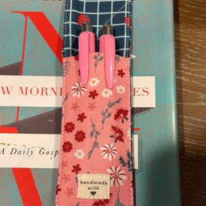 May include: A quilted fabric pen holder featuring a pink floral pattern on the front and a dark blue grid pattern with small strawberries on the top flap. Two pink pens are inserted into the holder, which has a small label reading handmade with a heart.
