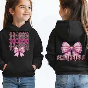 May include: A black hooded sweatshirt featuring the text JIU JITSU in a repeating pattern above a pink and white plaid bow on the front, and a large pink and white plaid bow above the text SMITHFIELD on the back.